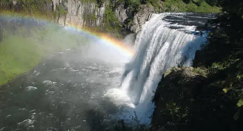 A large waterfall flows into a river with a visible rainbow in the mist.