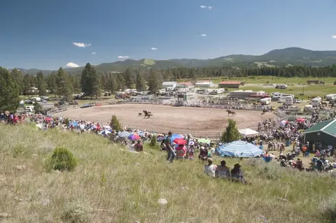 A wide outdoor shot of a rodeo arena with spectators gathered on a grassy hillside.