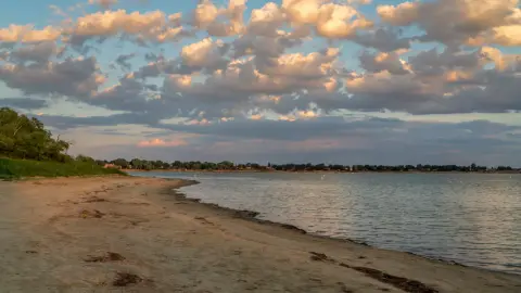 A sandy beach meets the water under a dramatic sky filled with clouds.