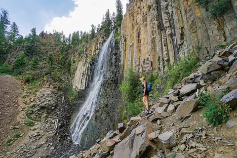 A hiker looks at a tall waterfall cascading down a cliff face with columnar rock formations.