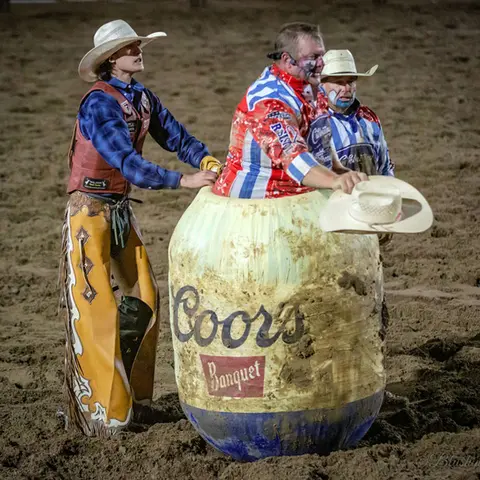 A rodeo clown inside a barrel with rodeo participants around him in a dirt arena.