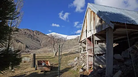 A weathered wooden building stands in a rural landscape with mountains in the background.