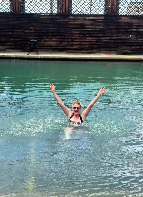 A woman splashing and smiling with her arms raised in a pool.