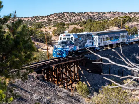 A decorated train crosses a wooden trestle bridge through a hilly landscape.
