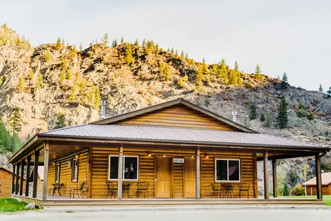 A log cabin building with a covered porch sits in front of a rocky hillside.