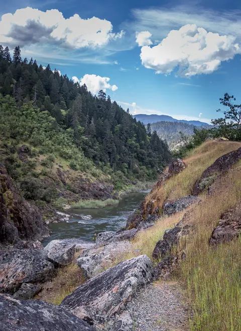 A river flows through a valley surrounded by forested hills and rocky grassy banks under a cloudy sky.