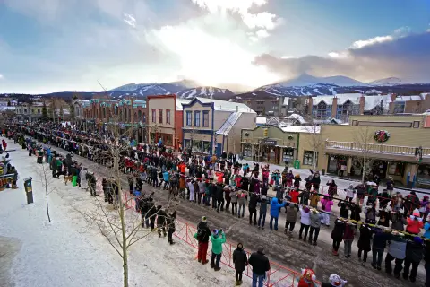 A large crowd gathers along a snowy street lined with historic buildings and mountains in the background.