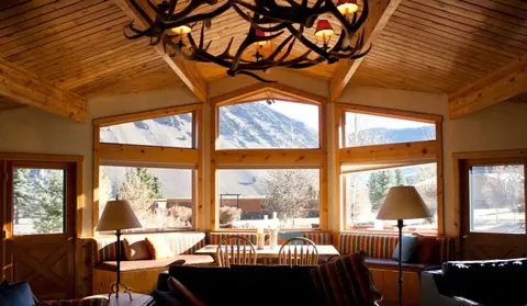 An interior shot of a rustic room with wooden paneling, large windows, and an antler chandelier.