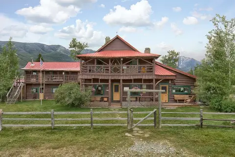 A large log cabin with a red roof stands behind a wooden fence with mountains in the background.