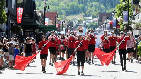 A marching band with red shirts and flags performs on a street lined with buildings and spectators.