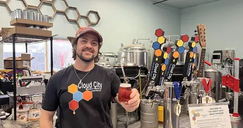 A man holds a glass of beverage inside a facility with brewing equipment and honeycomb decorations.