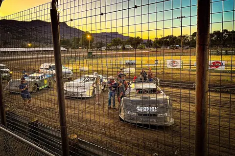 A view through a chain-link fence of race cars and people gathered on a dirt track at sunset.