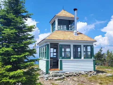 A small white and green fire lookout tower sits on a rocky mountain summit next to pine trees.