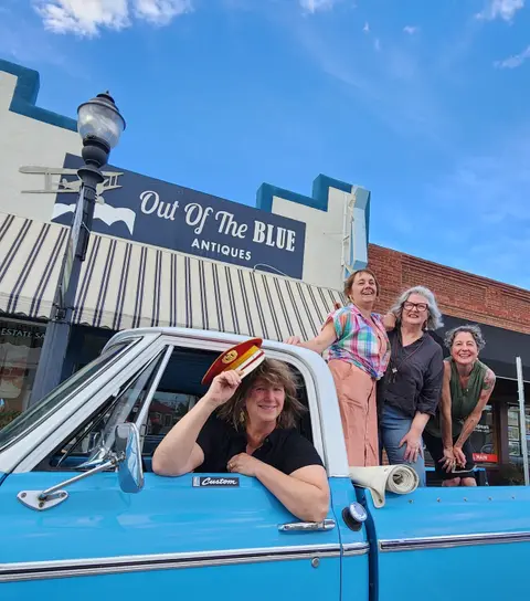 Four smiling women pose with a blue pickup truck parked on a street.