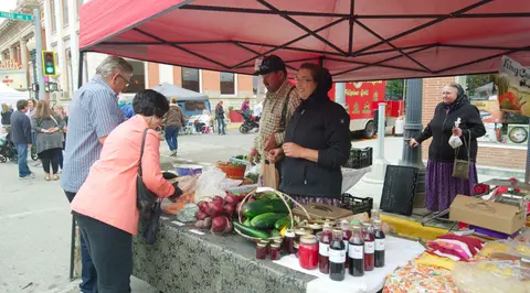 Vendors and customers interact at a farmers market stall under a red canopy.