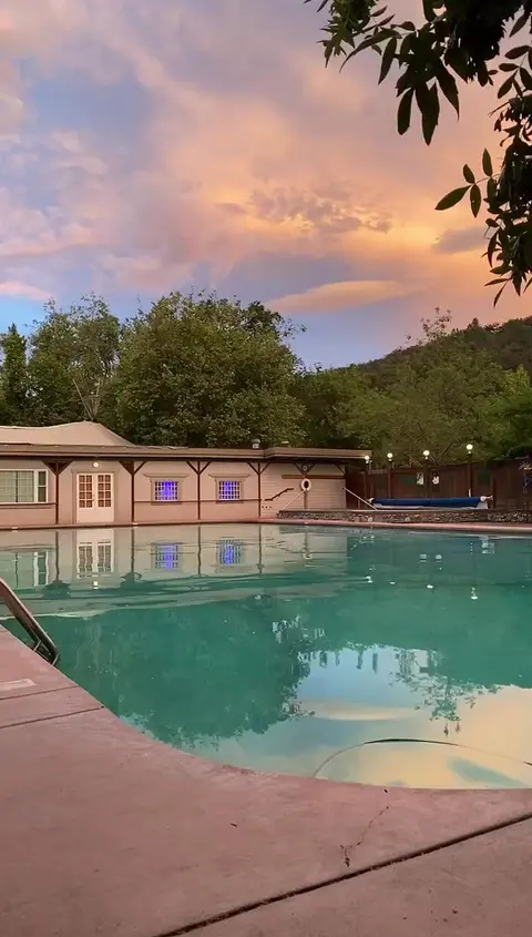 An outdoor swimming pool at sunset with a building and trees in the background.