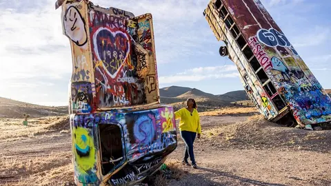 A person standing near colorful graffiti-covered stacked vehicles in a desert landscape.