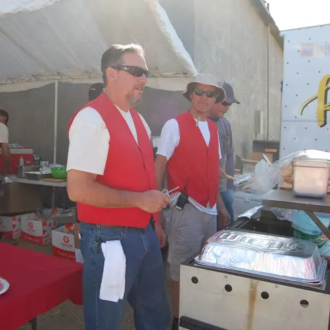 Two men wearing red vests stand by an outdoor food preparation station.