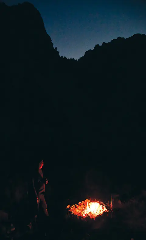 A person standing by a campfire at night with mountains in the background.