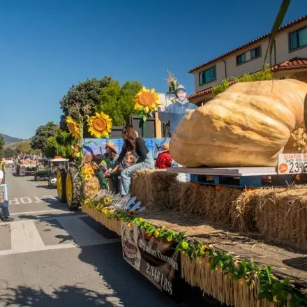 A parade float carries a giant pumpkin decorated with straw and sunflowers.