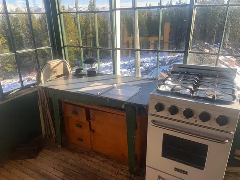 Interior of a cabin with a stove and table against large windows looking out to a forest and mountains.