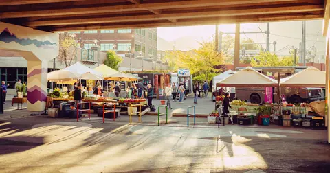 A bustling outdoor farmers market beneath an overpass with food trucks and tents.