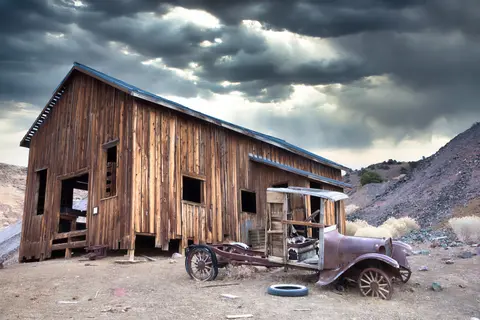 A dilapidated wooden building and a rusty vintage truck sit in a desert landscape under a stormy sky.