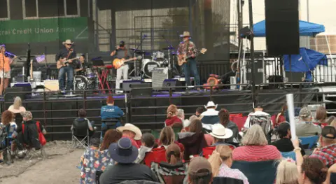 A band performs on an outdoor stage for an audience sitting in chairs.