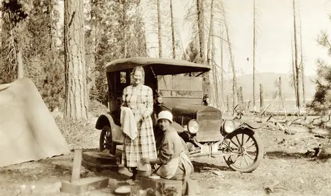Two women pose by a vintage car and tent in a wooded campsite.
