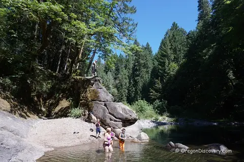 People swim and wade in a river next to a rocky cliff and forested banks.