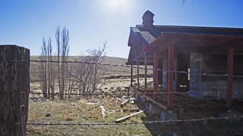 A rustic building stands in a dry landscape behind a barbed-wire fence.