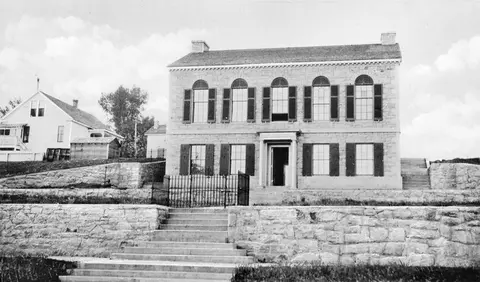 Historic black and white photograph of a large two-story stone building with a symmetrical facade and arched second-story windows.