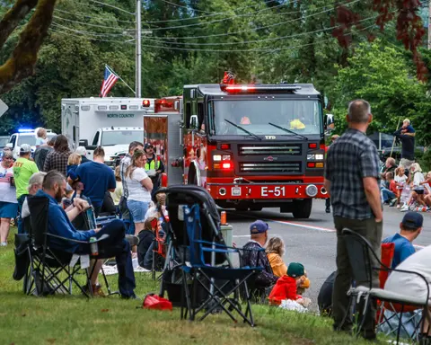 A fire truck drives down a street past a crowd of onlookers sitting in chairs.