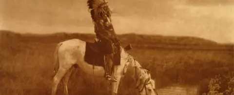 A historic sepia photograph of a person in a headdress riding a horse.