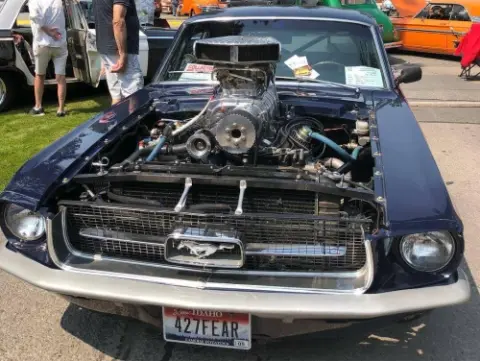 A classic car is parked outdoors at a vehicle show with its hood open to display a large engine.