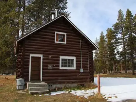 A brown log cabin stands surrounded by trees and patches of snow.