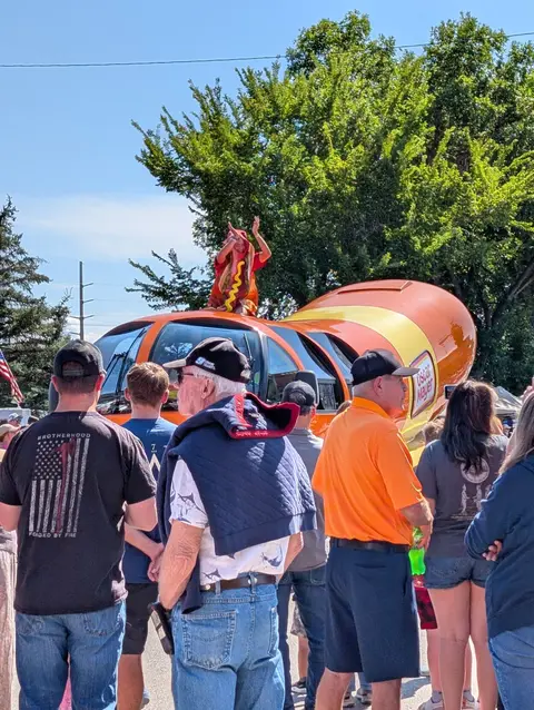 A crowd of people looks at a large hot dog vehicle outdoors.