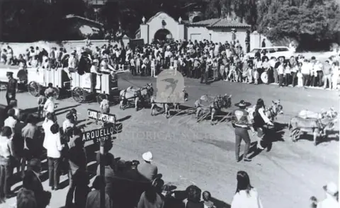 Black and white historic photo of a parade with horses, wagons, and a crowd.