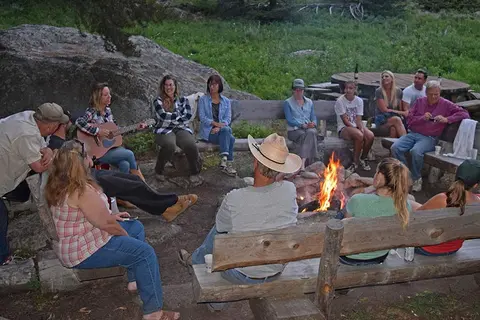 A group of people sitting around a campfire outdoors, with one person playing a guitar.