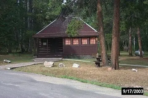 A rustic brown cabin sits among trees near a paved road.