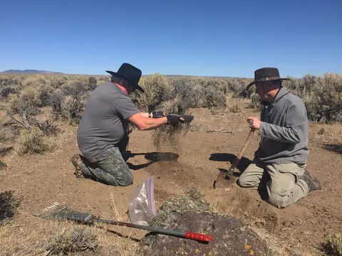 Two people digging and sifting soil outdoors in a dry landscape.