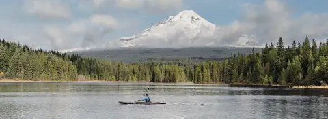 A person kayaking on a lake with a snow-covered mountain and forest in the background.