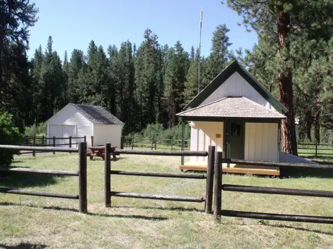 A small white building with a green trim stands in a grassy area with a wooden fence and a forest in the background.