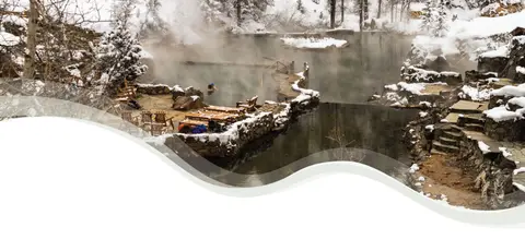 Steaming hot springs pool surrounded by snow and trees in winter.