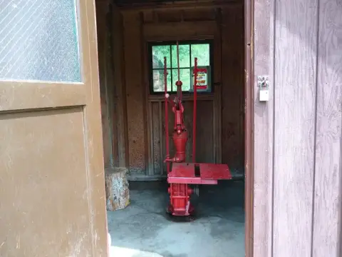 A red water pump stands inside a wooden shed viewed through an open doorway.