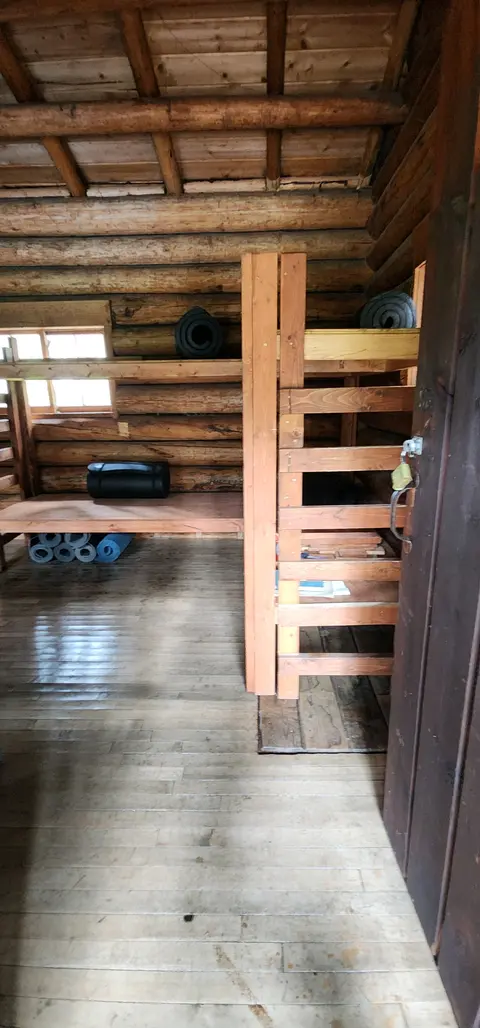An interior view of a rustic log cabin with wooden bunks and rolled sleeping mats.