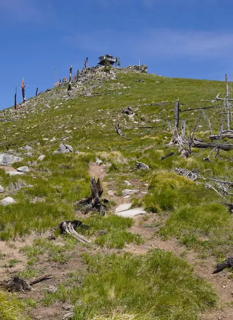 A fire lookout sits atop a grassy mountain peak with a blue sky above.