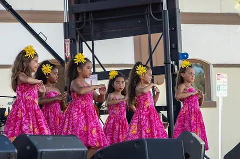 Six young girls in matching pink floral dresses and yellow hair flowers perform a dance on a stage.