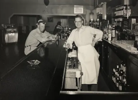 A historic black and white photograph shows a bartender standing behind a bar counter with a patron seated nearby.