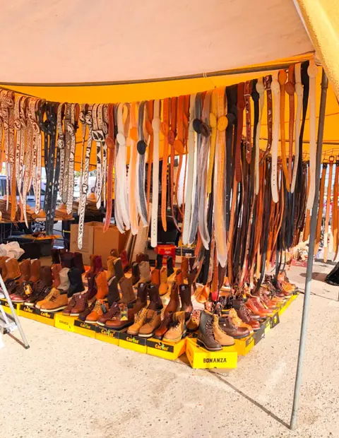 A vendor tent displays numerous leather boots and hanging belts for sale.